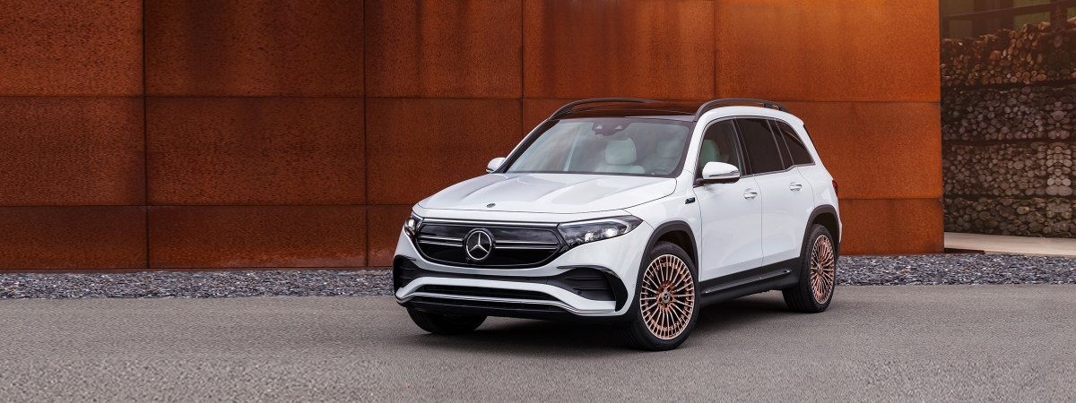 White Mercedes-Benz crossover SUV pictured in front of a rusty metal building, parked on light gray asphalt in front of a gravel border that reaches the rusty building. In the distance are neatly stacked cut lumber on industrial racks. The vehicle is a 2022 EQB, an electric version of the Mercedes GLB